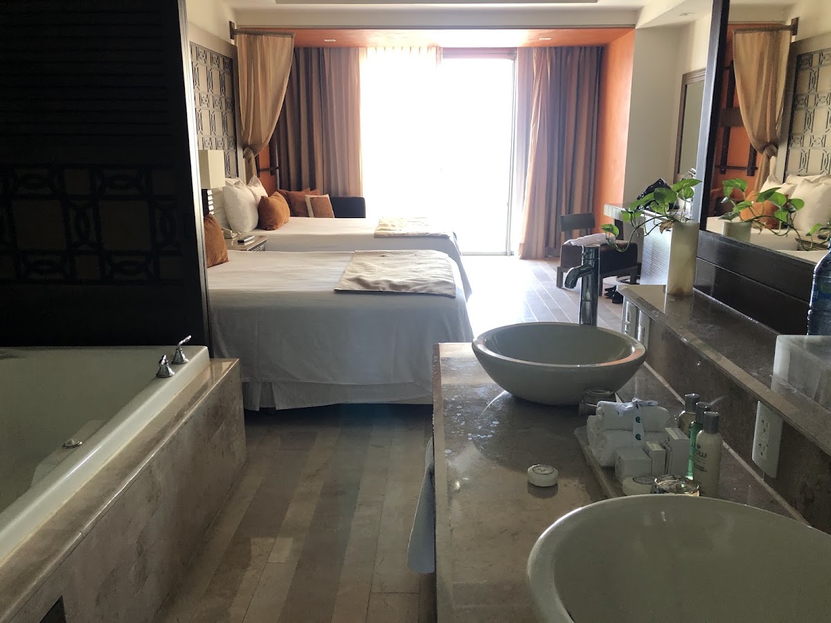 Resort suite bathroom with dual vessel sinks and soaking tub overlooking the bedroom with bed, seating area, and balcony doors with ocean views.