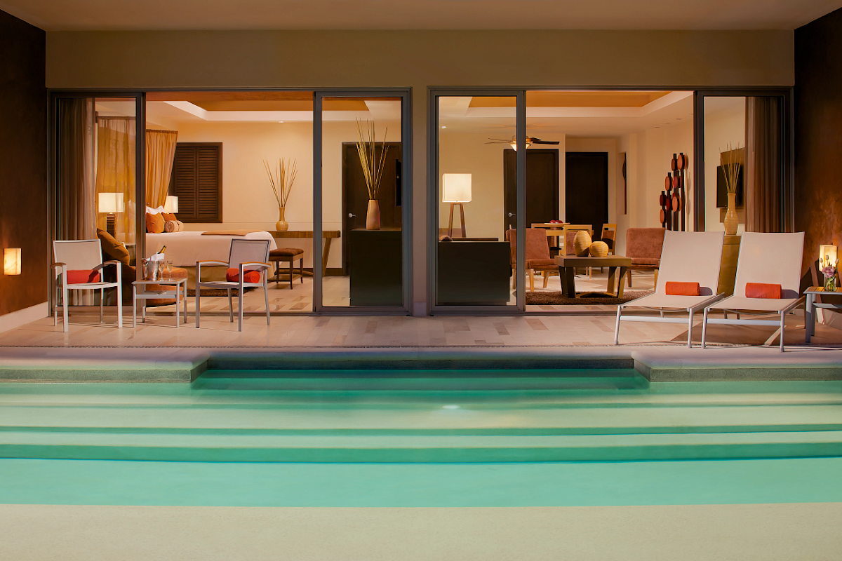 Dreams Vallarta Bay Resort & Spa suite with turquoise pool in foreground, illuminated interior visible through sliding glass doors with lounge chairs and furnishings.