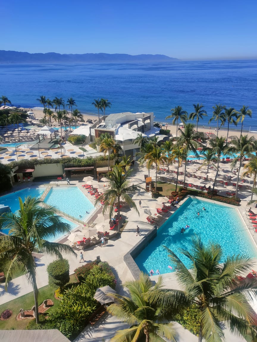 Aerial view of Dreams Vallarta Bay Resort & Spa with multiple pools, palm trees, sandy beach, and blue ocean with mountains in the distance.