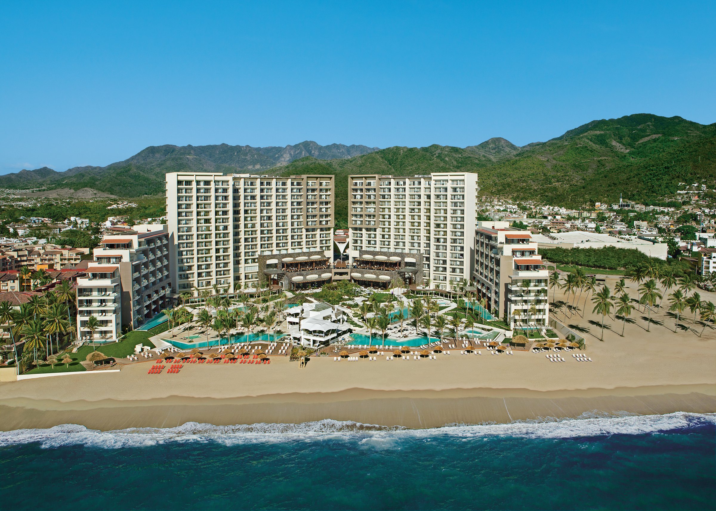 Wide aerial of the U-shaped resort, its pools and the beach with the bay and mountains.