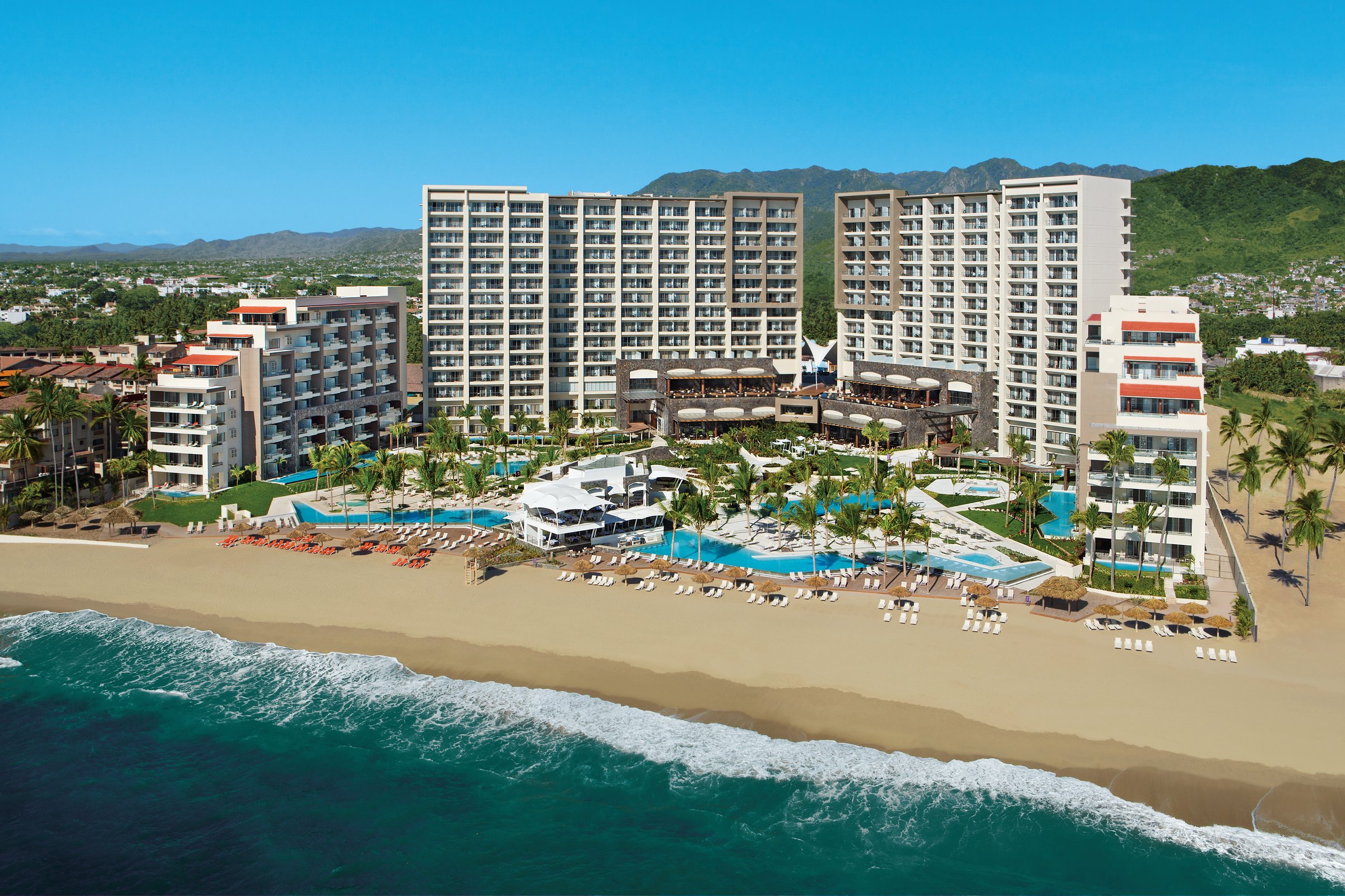 Closer aerial of the glass-balconied guest buildings, pools and beach loungers.