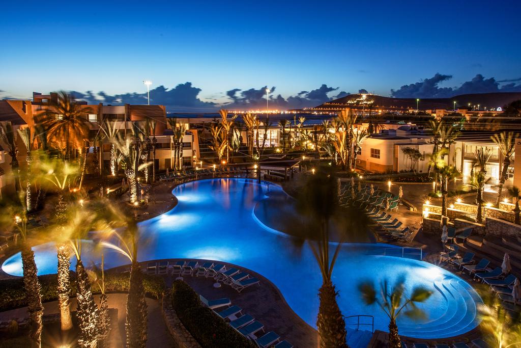 Illuminated pool complex at dusk with blue water, palm trees, lounge chairs, and beachfront buildings at Dunes d'Or Ocean Club.
