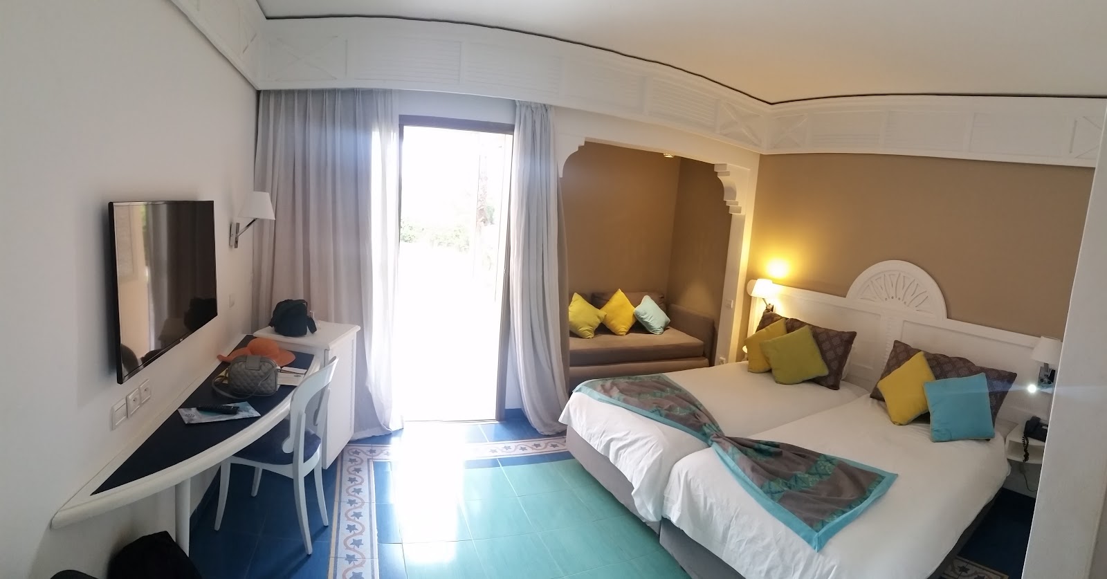 Resort bedroom with two beds featuring patterned throws, yellow and brown decorative pillows, warm beige walls, and sheer curtains framing a window.