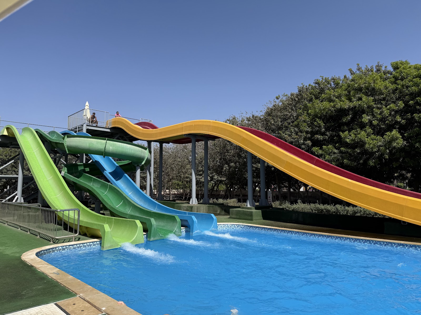 Water slides in bright green, blue, and yellow-red curves descend into a blue pool at Dunes d'Or Ocean Club, with trees and clear sky beyond.