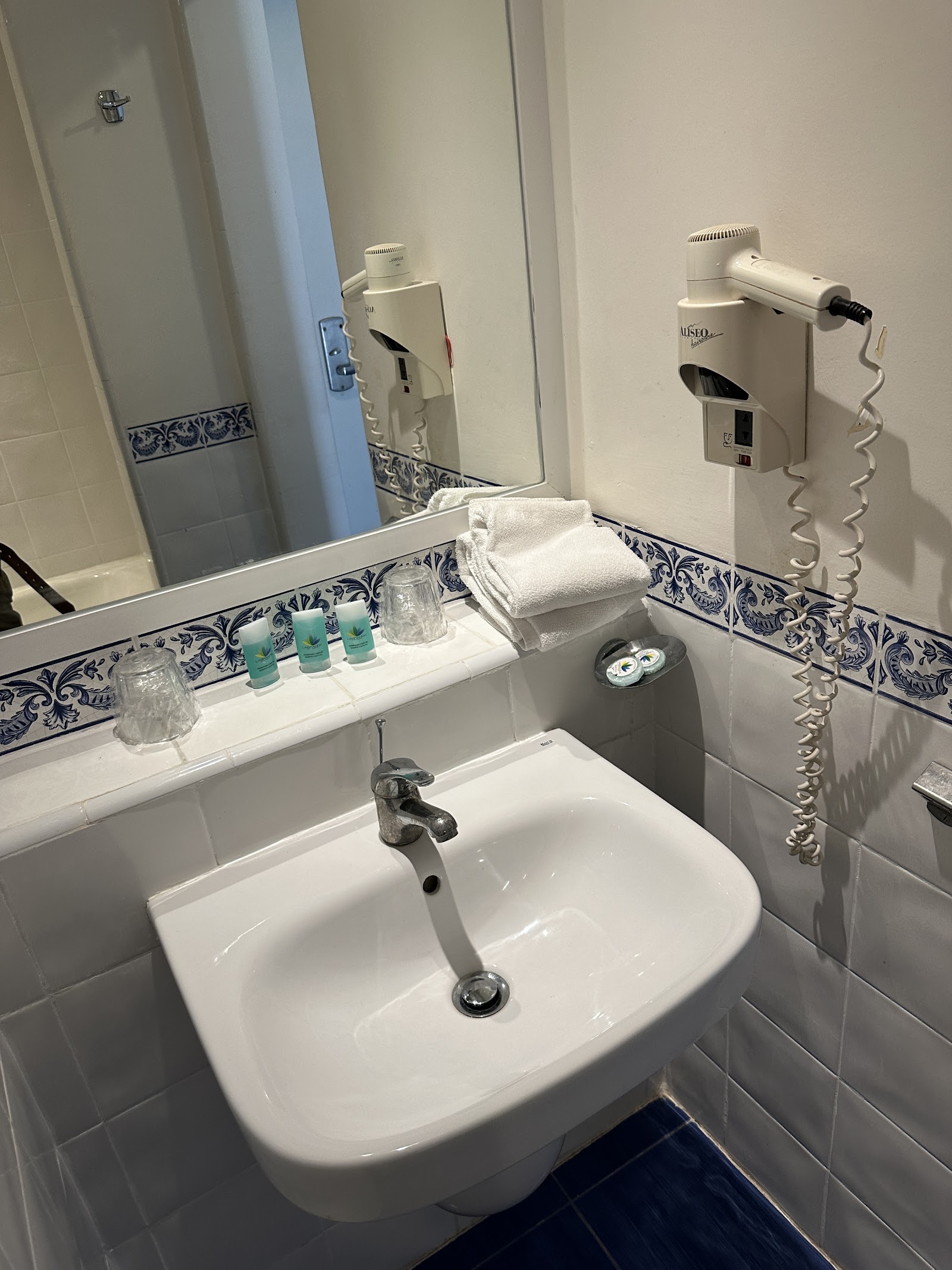 Clean white bathroom with blue patterned tile trim, white sink, mirror, toiletries, towels, and wall-mounted hair dryer at Dunes d'Or Ocean Club.