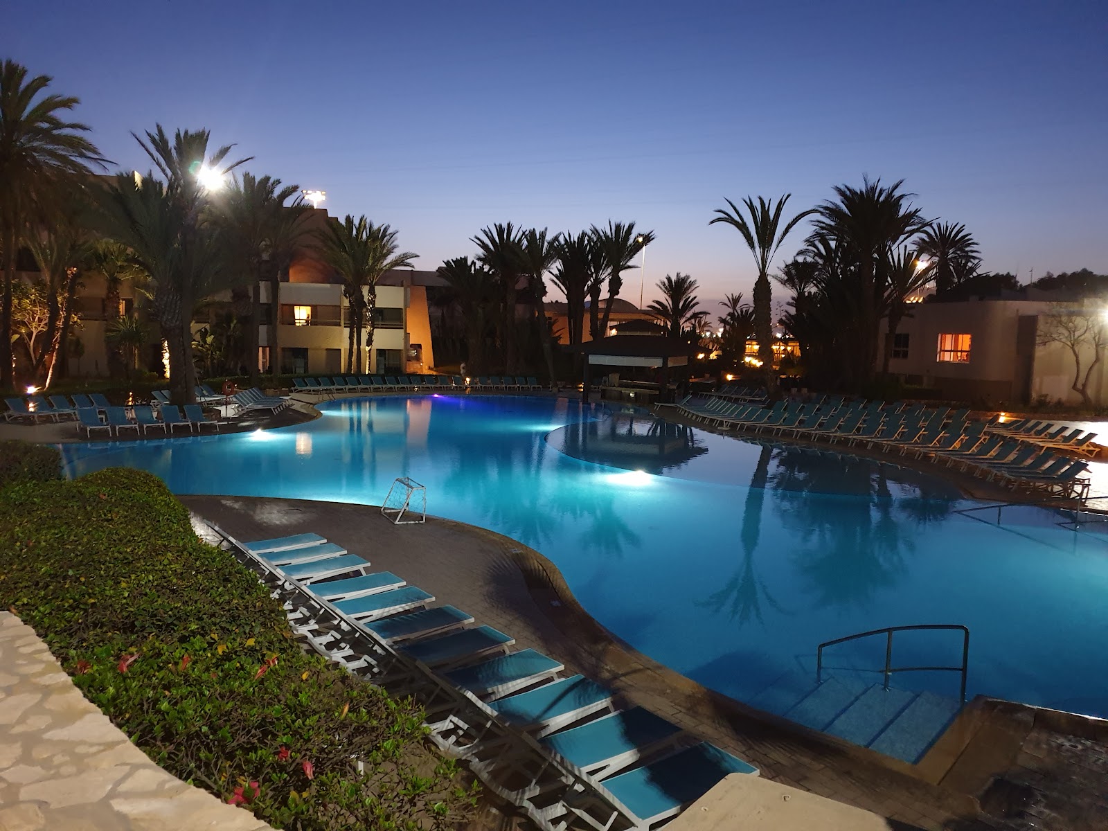 Illuminated lagoon-style pool surrounded by blue loungers, palm trees, and lit resort buildings at dusk.