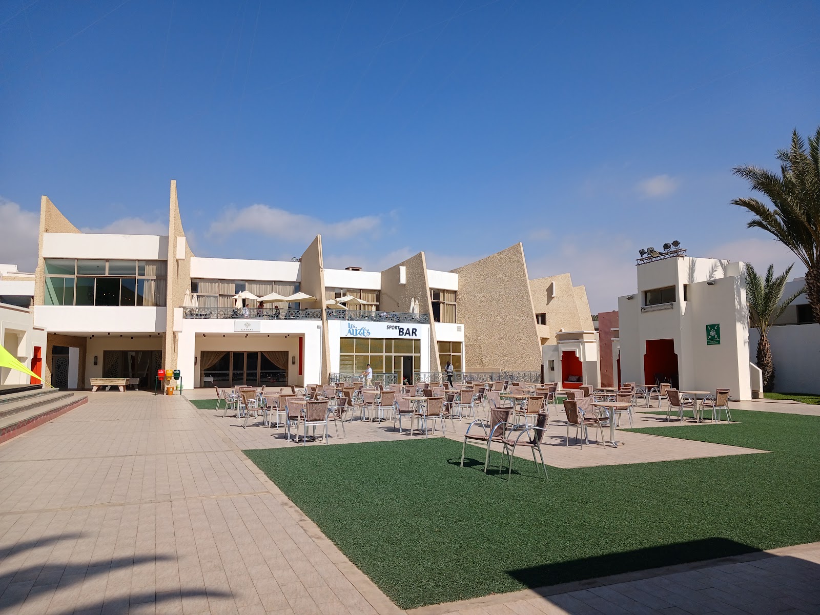 Central courtyard with metal chairs scattered on green lawn and paved areas, bordered by geometric white and tan resort buildings under clear blue sky.
