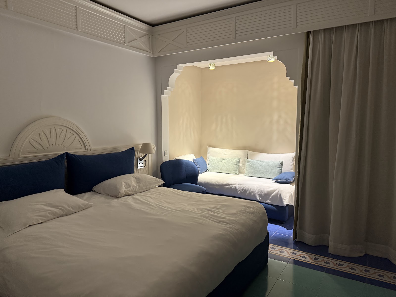 Two double beds with white linens decorated with blue towel bundles tied with red flowers in a cream-colored resort bedroom with wall sconces and a white headboard.