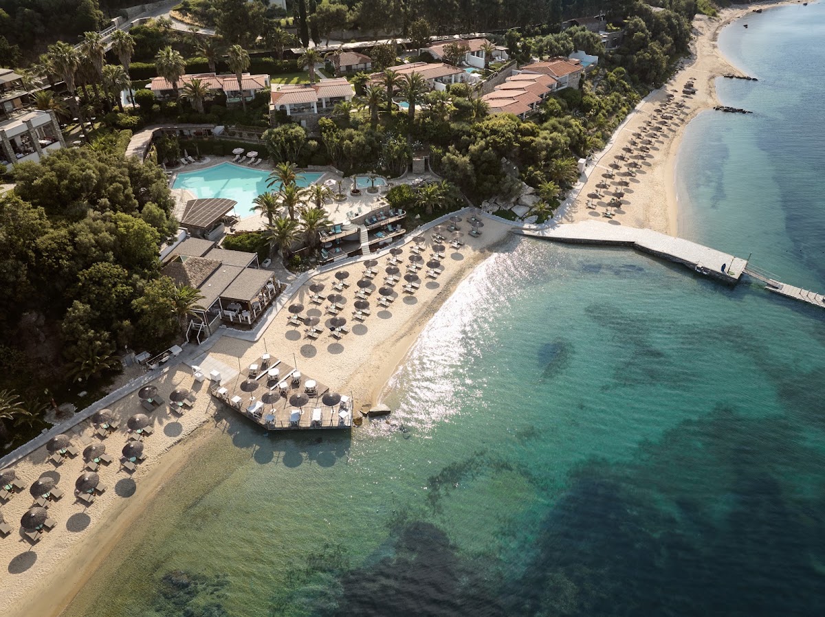 Eagles Palace beachfront with sunbed-lined sandy shore, turquoise water, pool, and Mediterranean buildings nestled among pine trees.