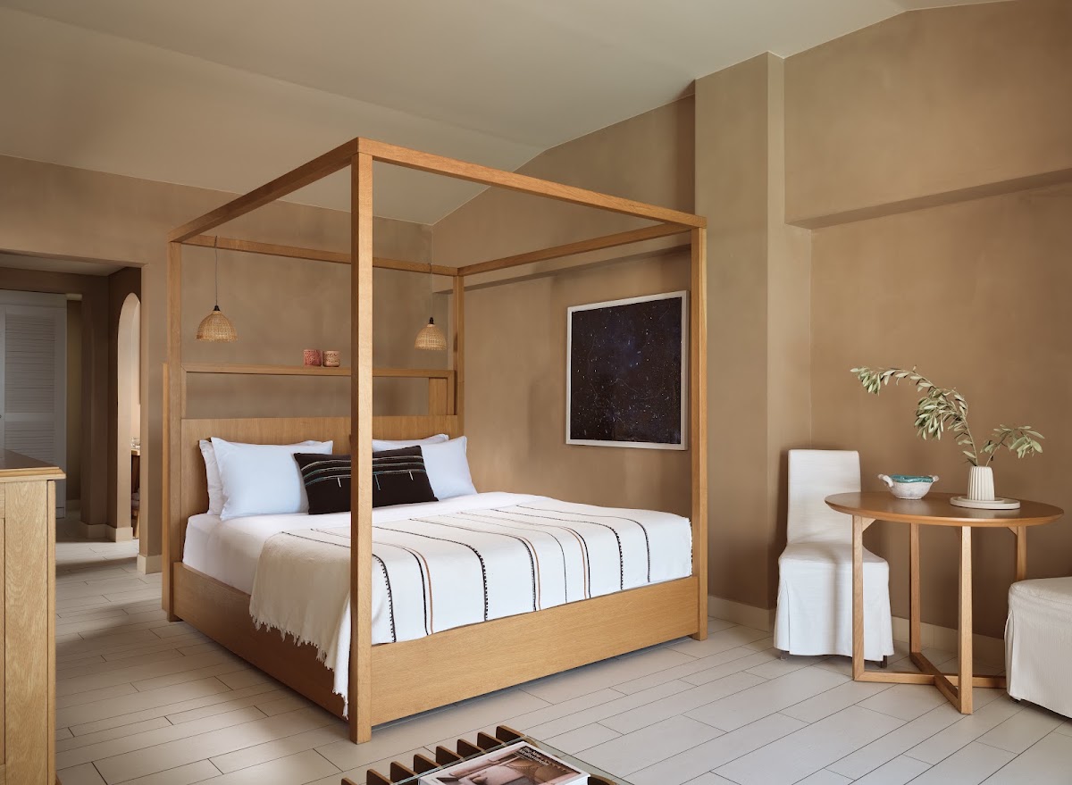 Spacious bedroom with wooden four-poster bed, white bedding, side table with potted plant, and neutral beige walls throughout.