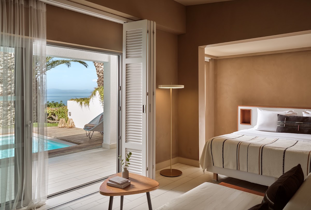 Resort bedroom with ocean view through open glass doors onto a terrace with lounge chair and sea beyond.