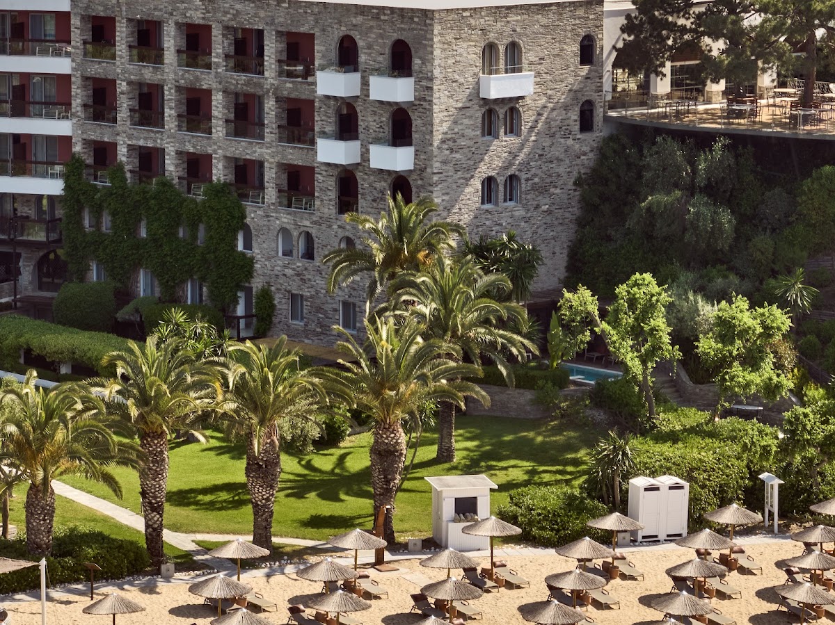 Stone hotel building with red-shuttered windows overlooking landscaped gardens with palm trees, beach area with thatched umbrellas and loungers in foreground.