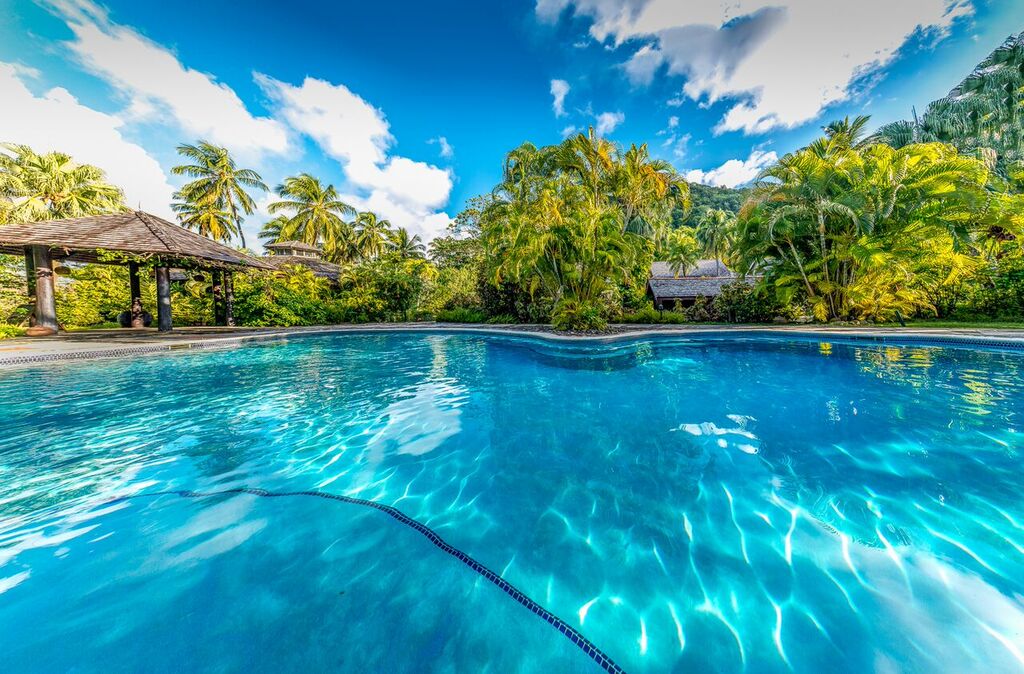 Clear blue lap pool with lane divider, wooden pergola, and lush tropical vegetation surrounding East Winds resort under bright sky.