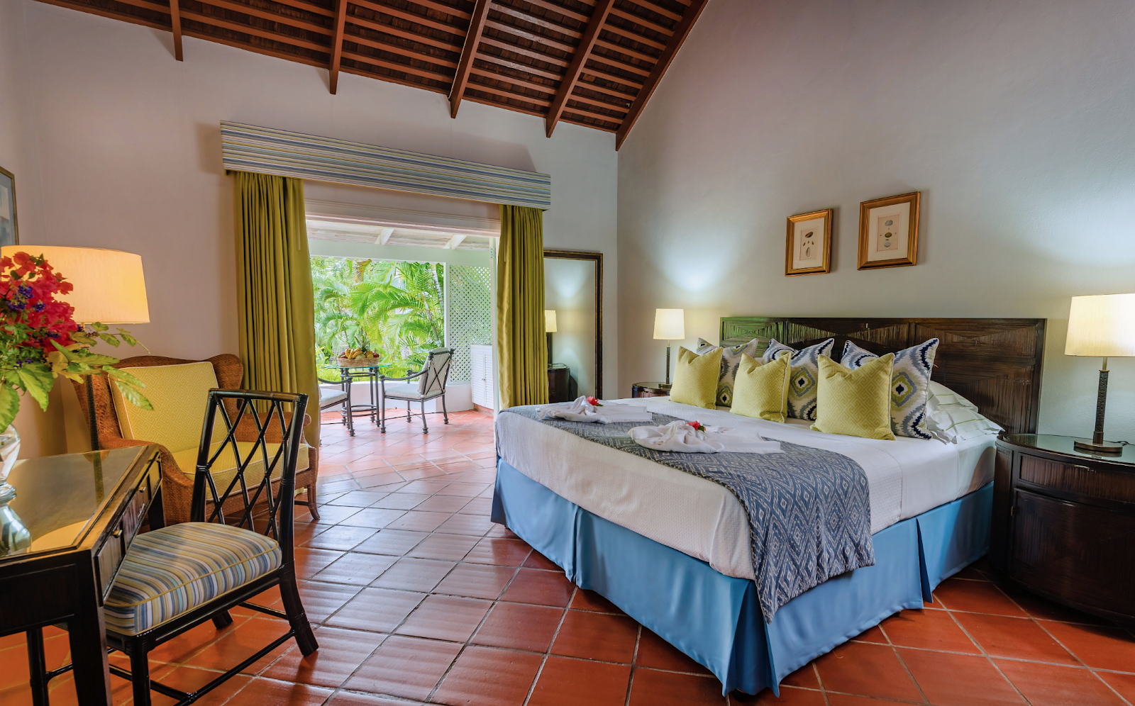 Spacious bedroom at East Winds with king bed, yellow and blue accents, terracotta tile floor, and garden view through French doors.