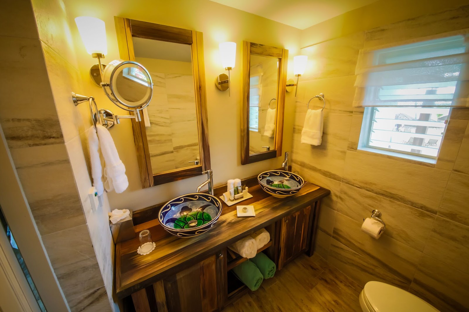 Bathroom with dual wooden vanity, decorative ceramic sinks, illuminated mirrors, and warm yellow tile walls at East Winds.