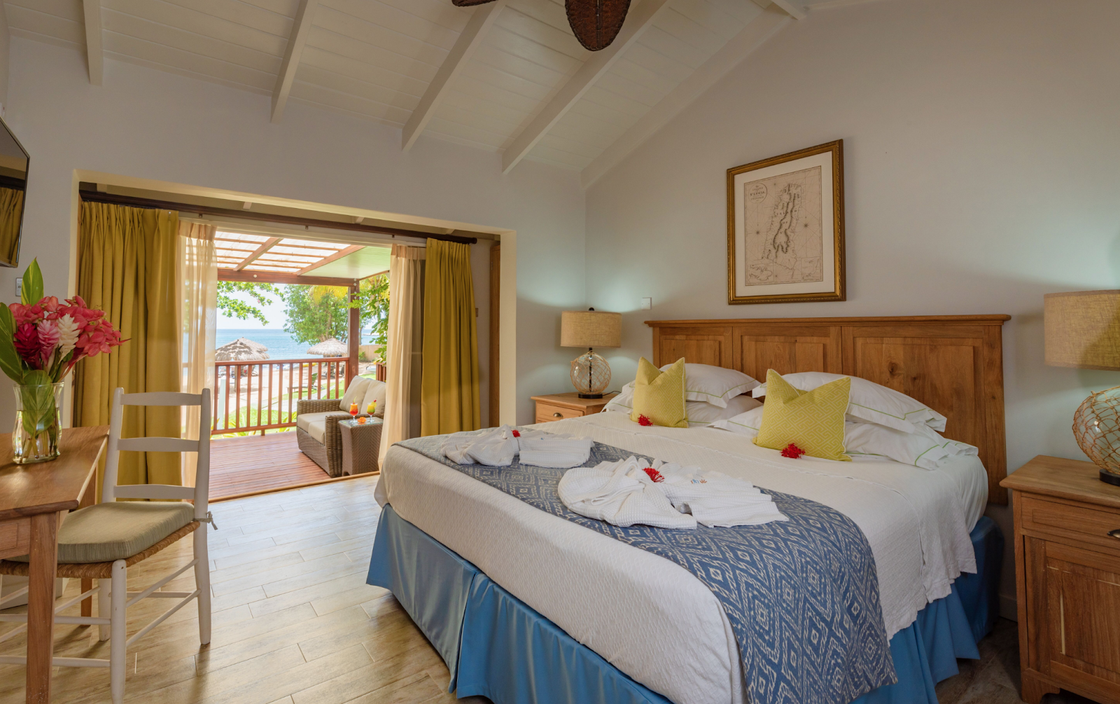 Resort bedroom with wooden bed, yellow pillows, ocean view through glass doors opening to a balcony with sea visible beyond.