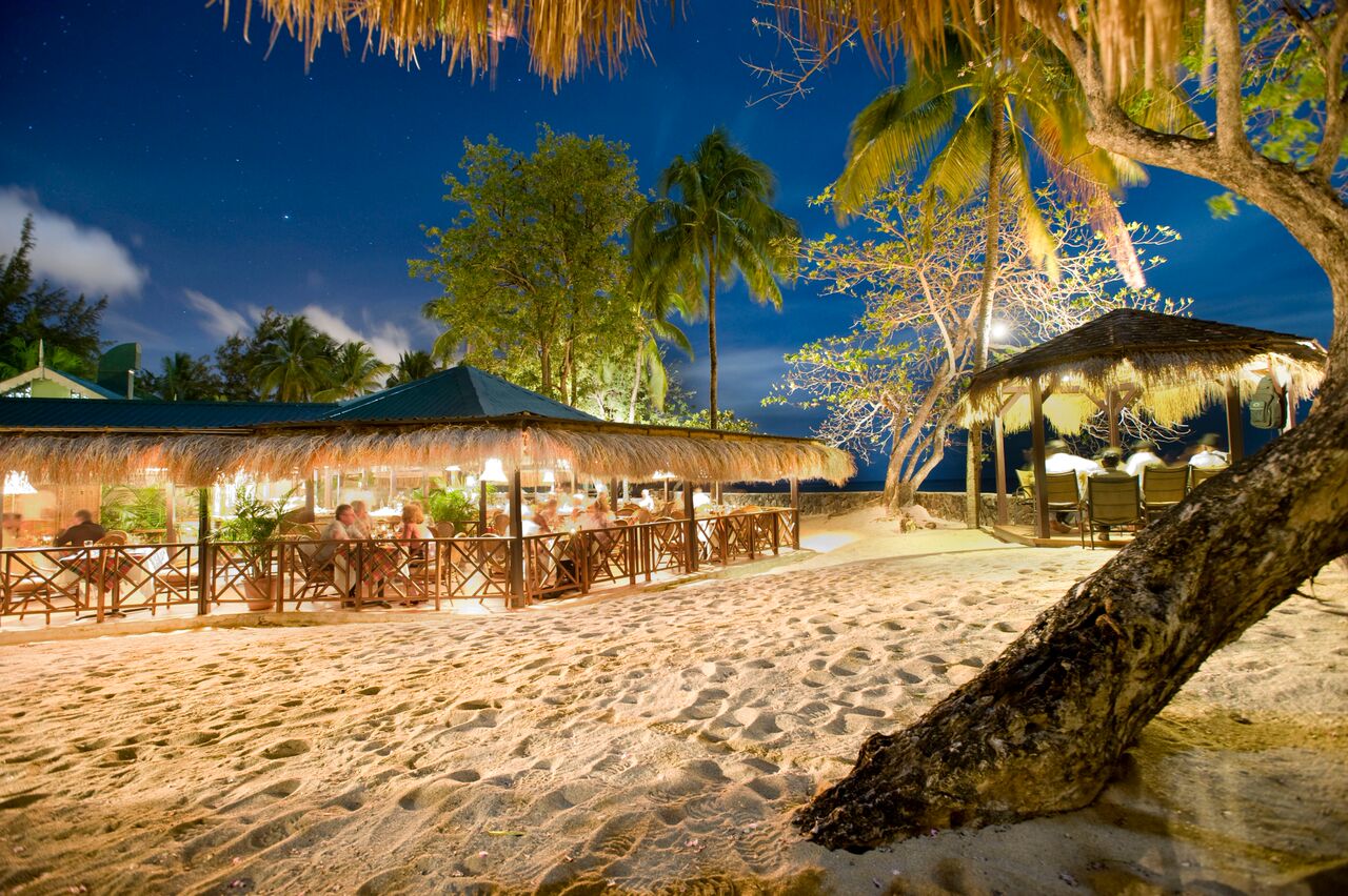Beach dining area at East Winds with thatched-roof restaurants, wooden chairs, palm trees, and dark ocean under twilight sky.