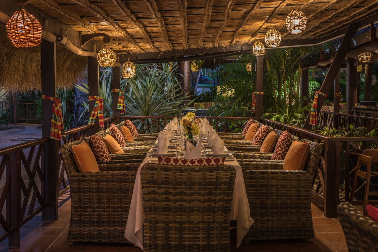 Long dining table set for dinner under thatched roof with woven lanterns, tropical plants, and ocean view at East Winds.