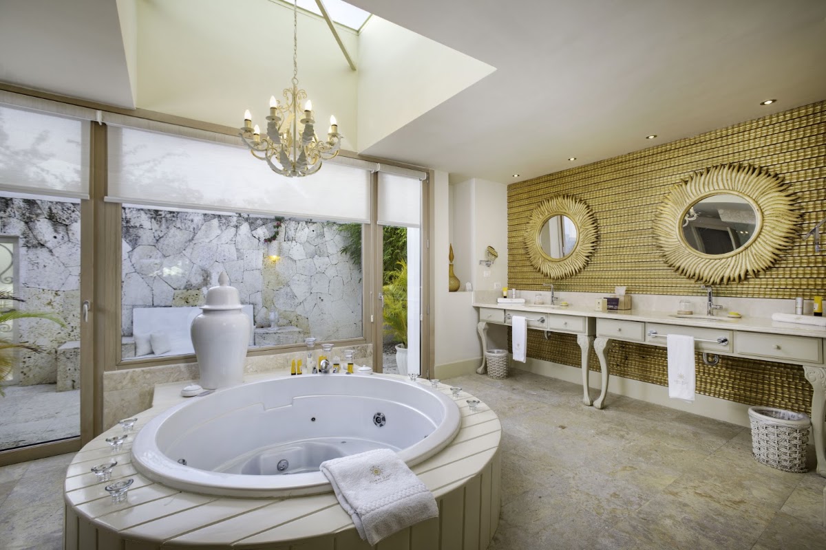 Spacious bathroom with sunken circular soaking tub, dual vanities with gold-framed mirrors, chandelier, and views of stone courtyard at Eden Roc Cap Cana.