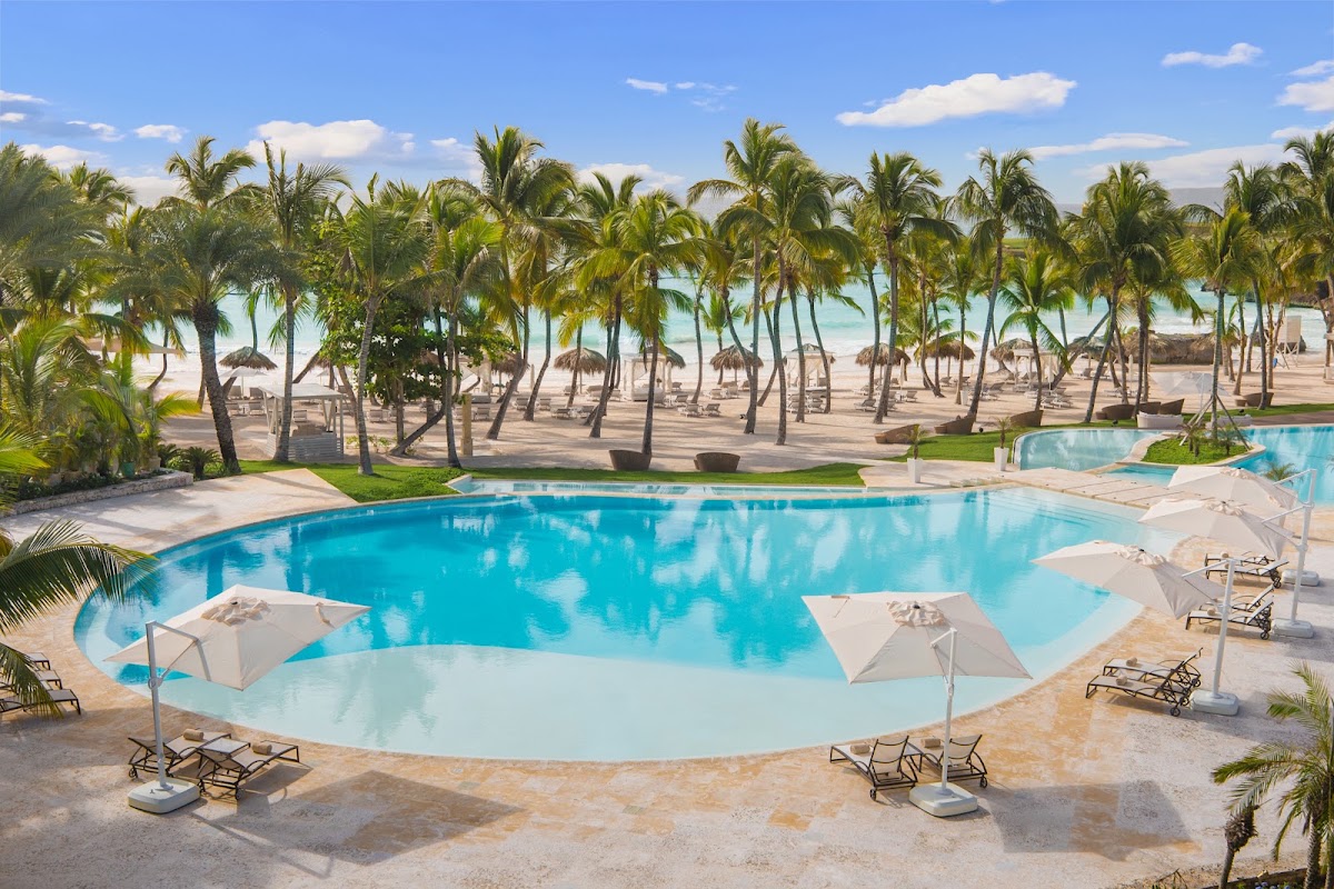 Resort pool with lounge chairs and white umbrellas overlooking palm-lined beach and turquoise ocean at Eden Roc Cap Cana.