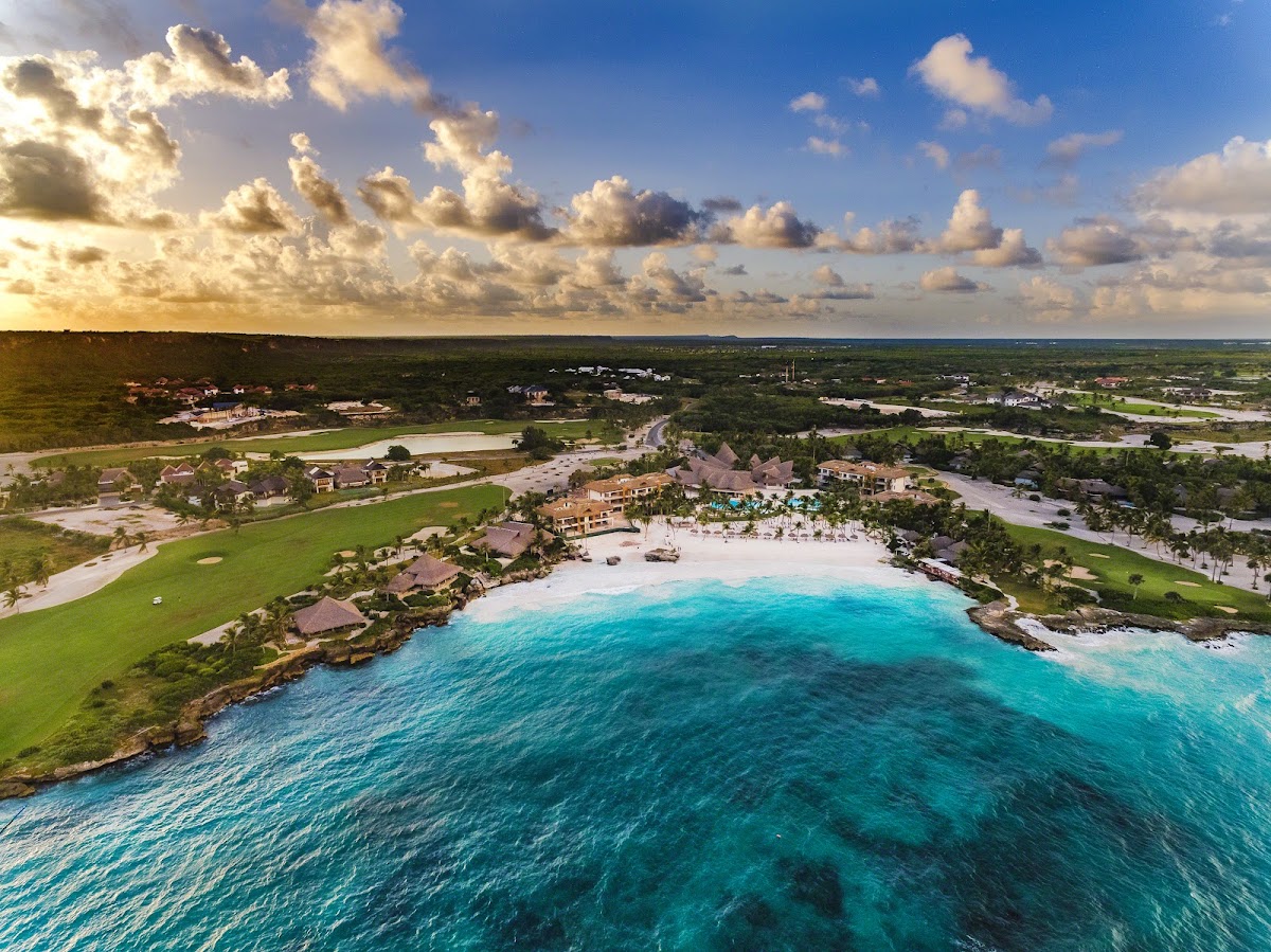 Aerial view of Eden Roc Cap Cana beachfront resort with turquoise water, white sand beach, thatched-roof buildings, and green grounds along the Dominican coast.