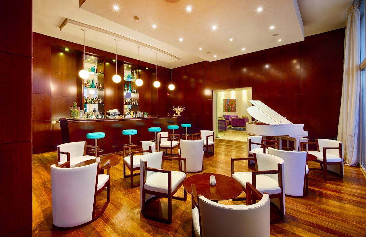 Upscale lounge with deep red walls, white curved seating, turquoise bar stools, wooden floors, and a white grand piano.