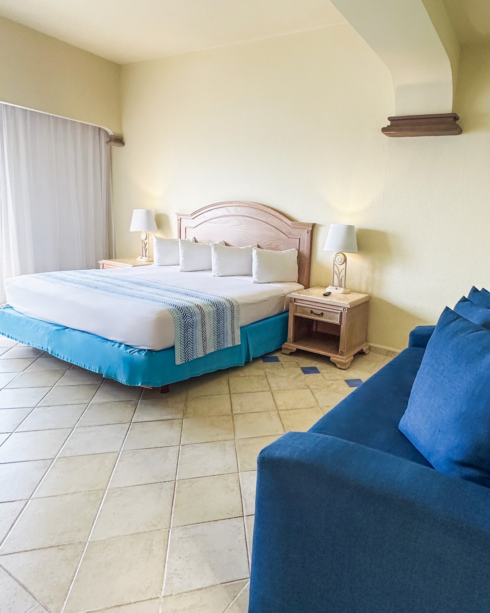 Resort bedroom with queen bed in turquoise skirt, white pillows, wooden headboard, blue armchair in foreground, and tile flooring.