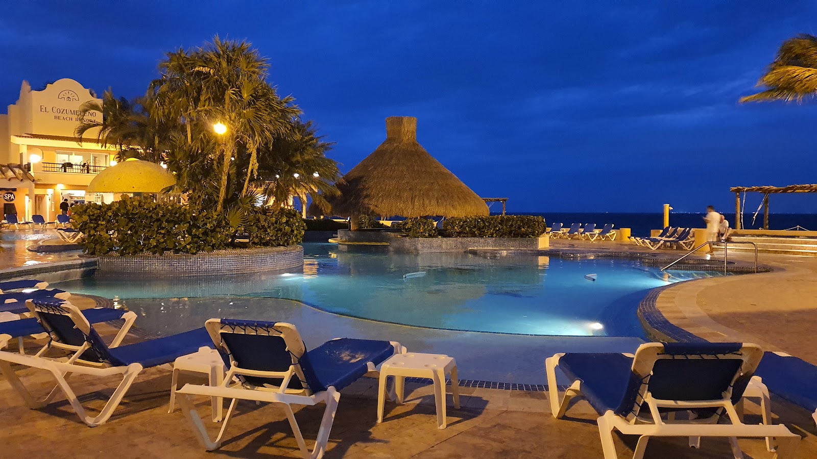 Lit pool at night surrounded by lounge chairs and palm trees, with ocean and beach bar visible in background.