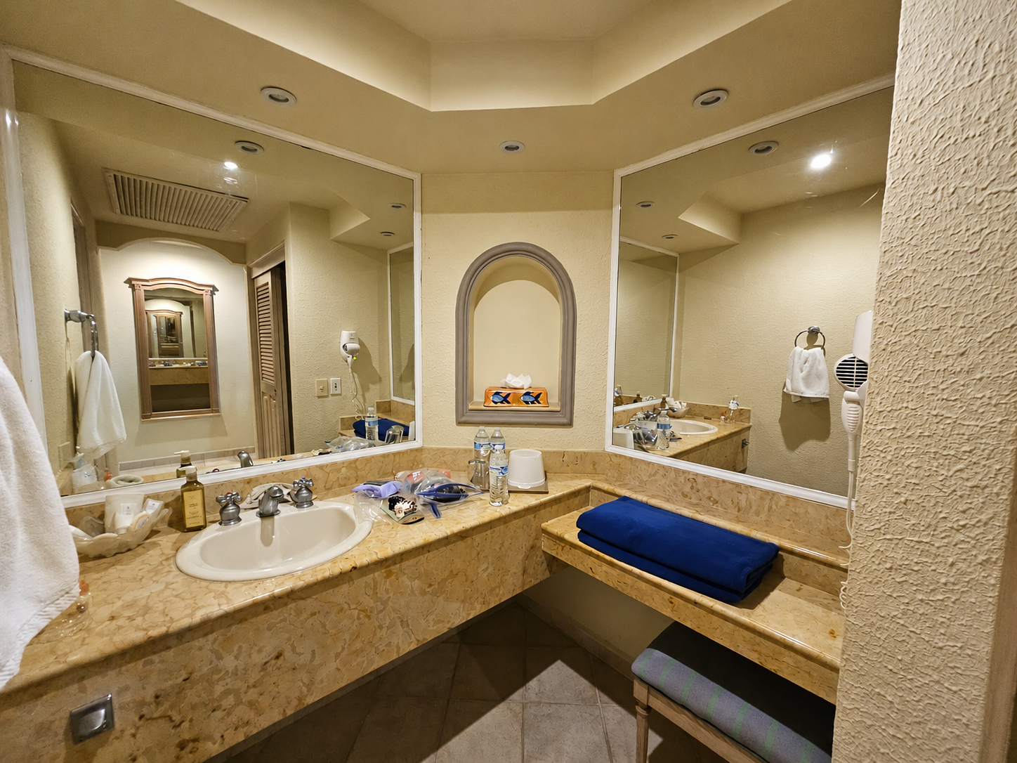 Spacious bathroom with marble countertop, dual sinks, large mirrors, and shelving with rolled towels.