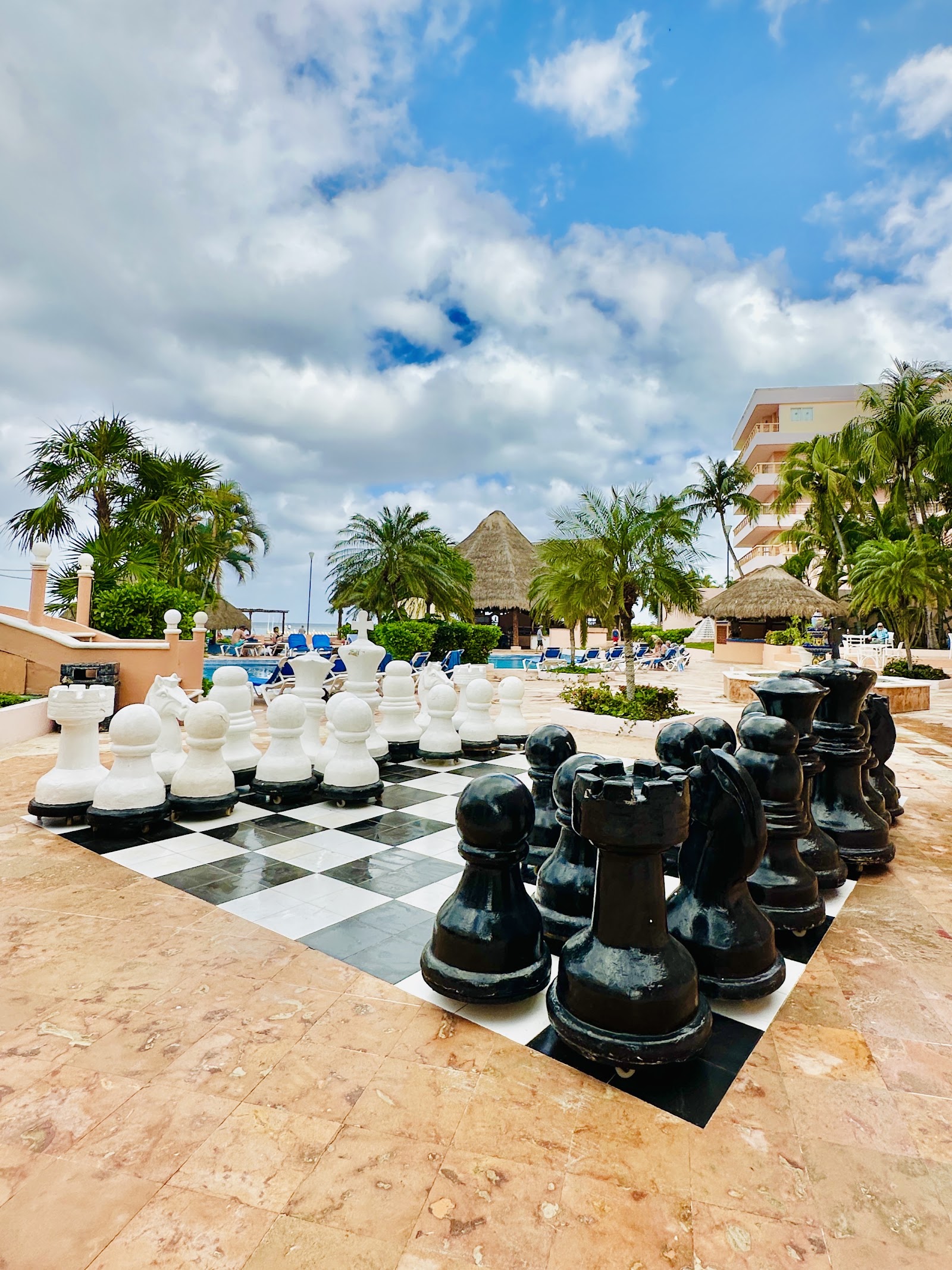 Oversized chess game on a tiled patio at El Cozumeleño Beach Resort, with thatched-roof structures and palm trees in the background.