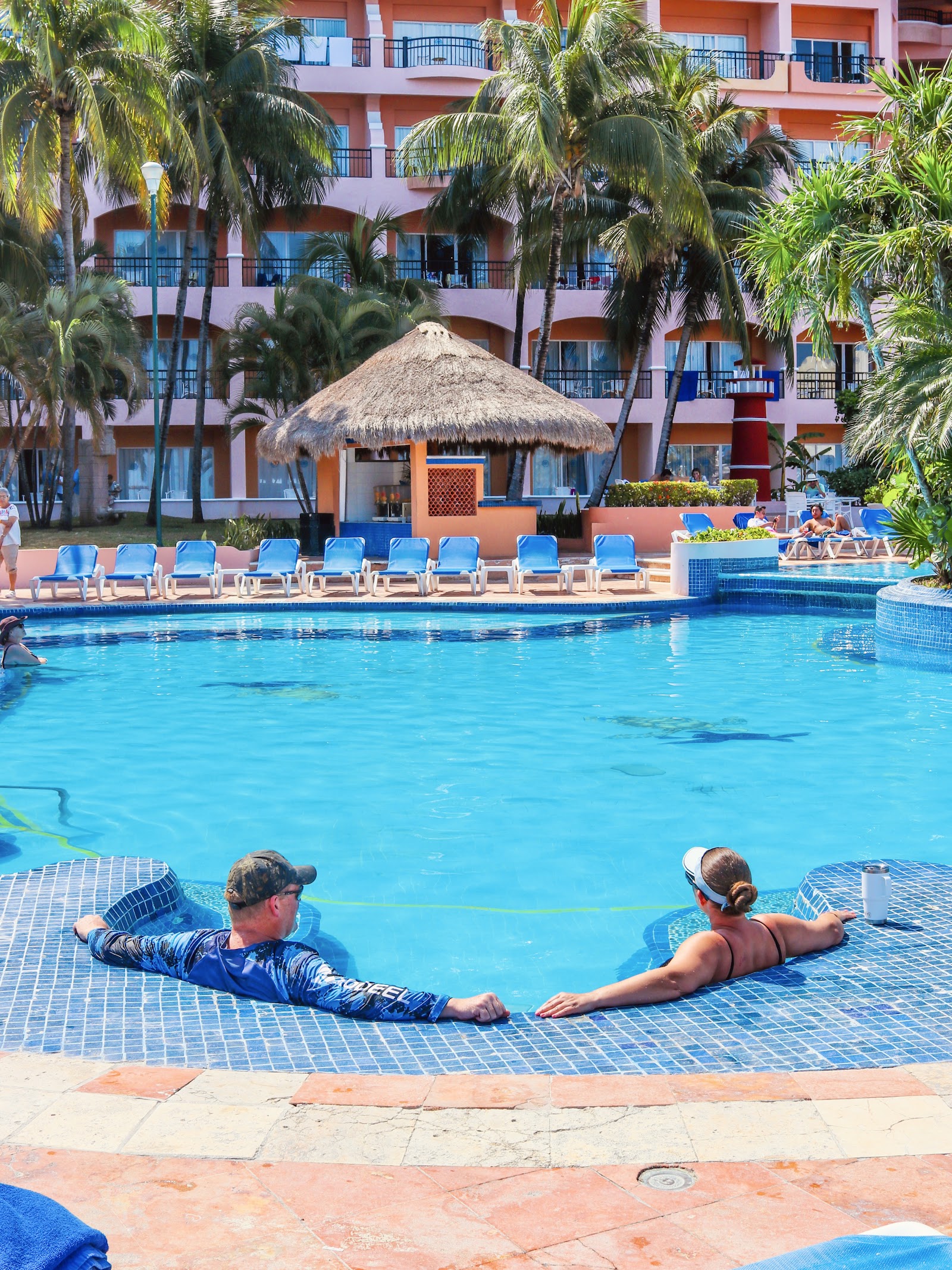 Blue loungers line the beach and pool deck at El Cozumeleño, with thatched palapas, turquoise water, and palm trees visible beyond.