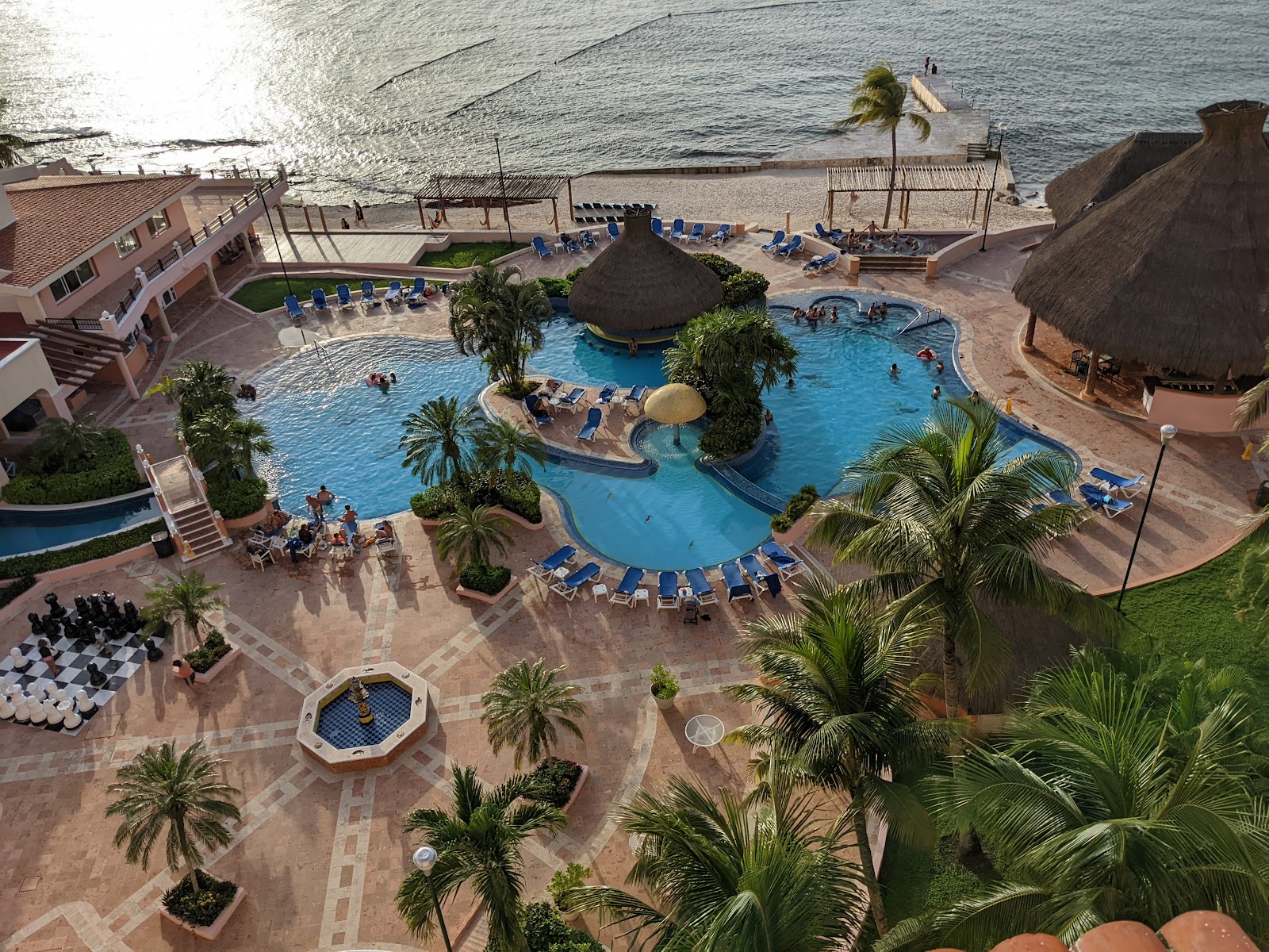 Indoor thermal pool with light blue water surrounded by large limestone rocks, palm trees, and the orange and white building structure of El Cozumeleño Beach Resort.