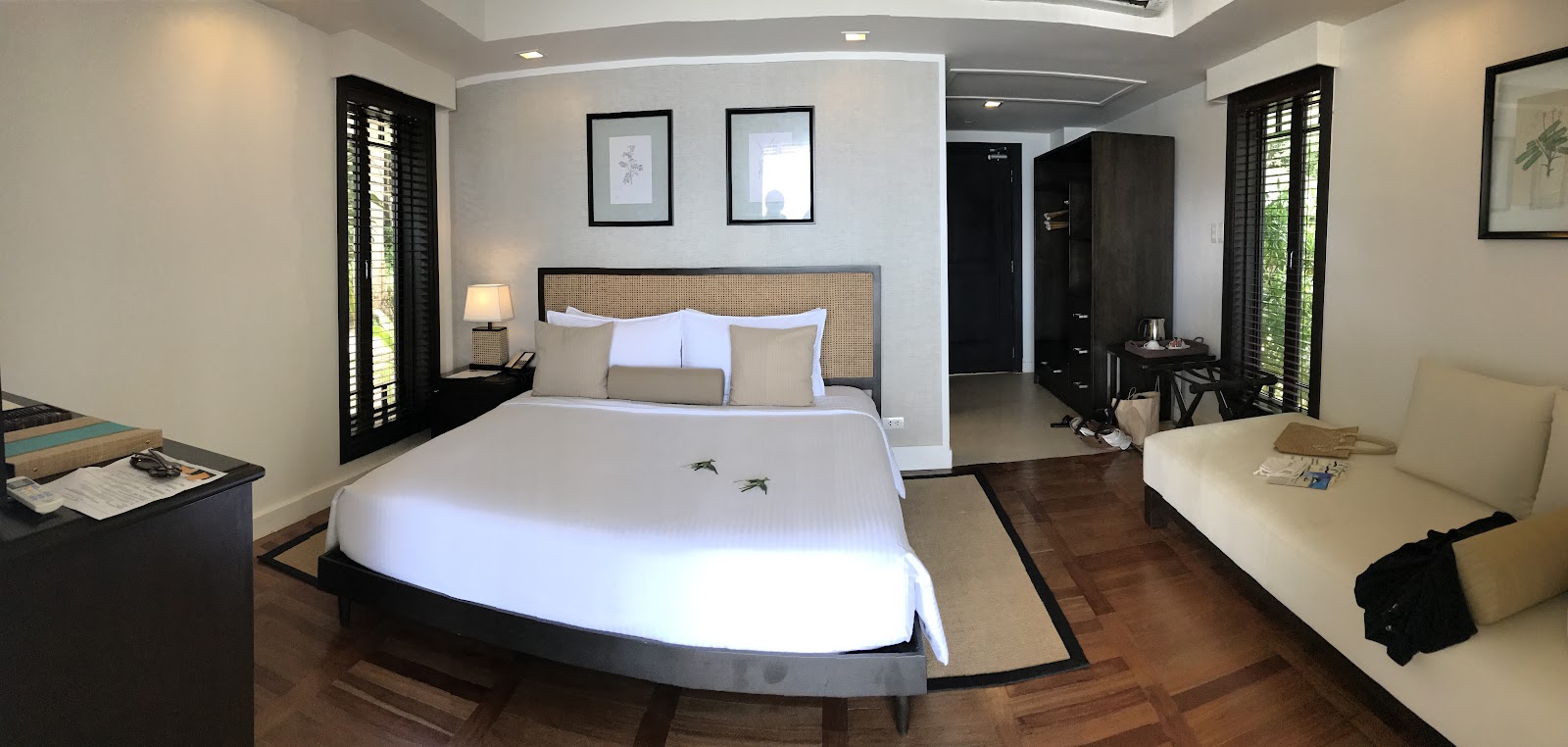 Resort bedroom with curved bed, rattan headboard, cream sofa, dark wood floors, and black shuttered doors on either side.