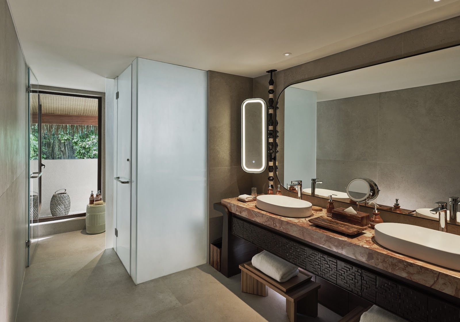 Modern bathroom with dual vessel sinks on wooden counter, illuminated mirror, and walk-in shower opening to outdoor garden area.