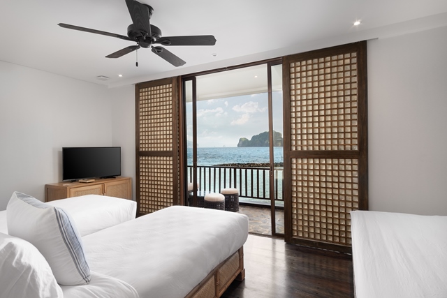 Hotel room with two beds, wooden TV stand, and lattice doors opening to a balcony overlooking tropical limestone cliffs and sea.