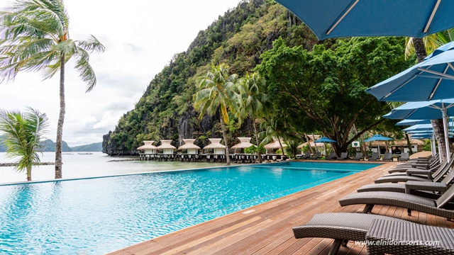 Beachfront pool with palm trees, thatched-roof bungalows, and limestone cliffs across turquoise water at El Nido Resorts Miniloc Island.