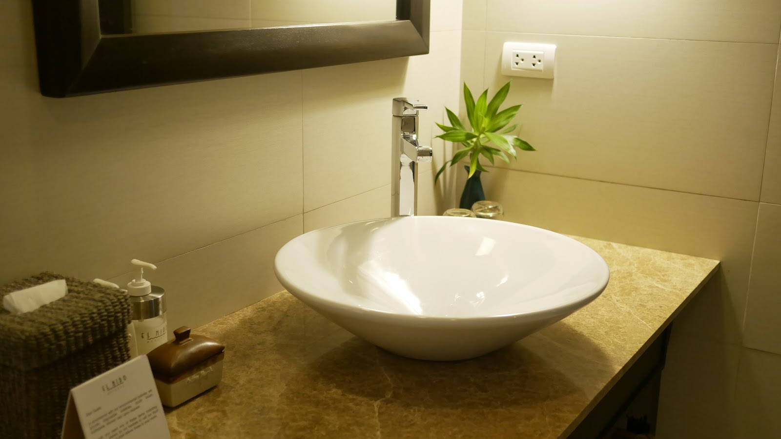 Modern bathroom with round white vessel sink on stone countertop, tall faucet, mirror, and green plant in black vase.