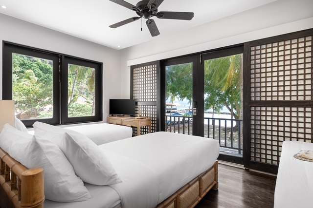 Bright beachfront bedroom with two beds, black-framed doors opening to ocean views and palm trees, dark wood flooring, and ceiling fan.