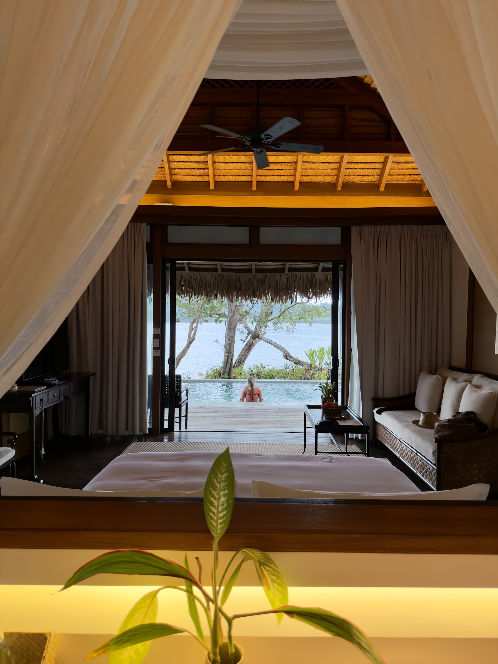 Overwater bungalow interior framed by sheer curtains, with bed, ceiling fan, and open doors revealing a lagoon, trees, and a guest in the water beyond.