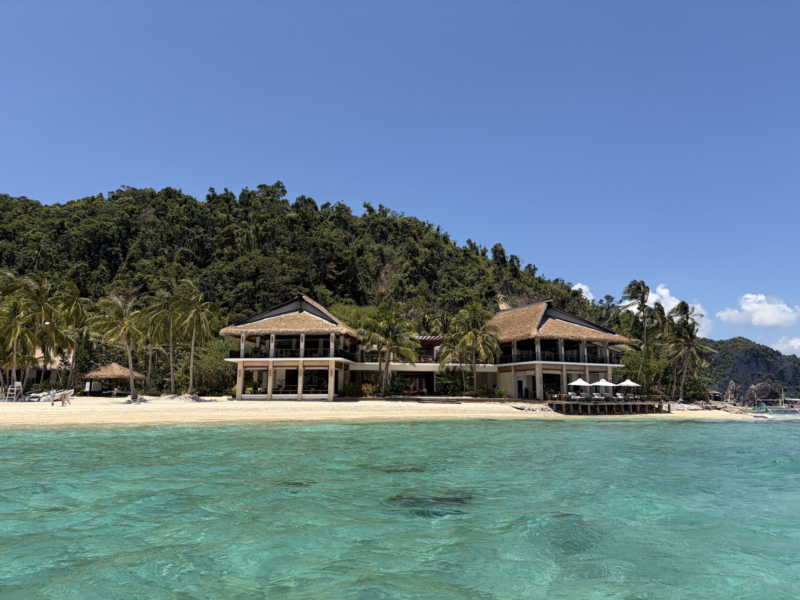 Two beachfront buildings with thatched roofs sit on white sand backed by dense tropical forest, viewed from clear turquoise water.