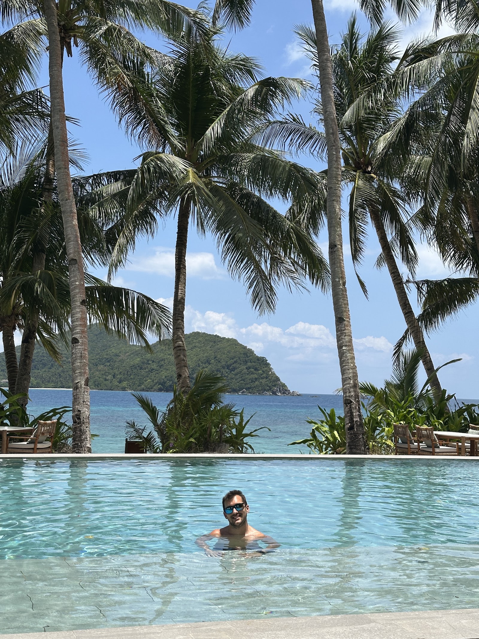 Man in sunglasses swimming in shallow lagoon pool bordered by tall palm trees, with forested island and turquoise ocean beyond.