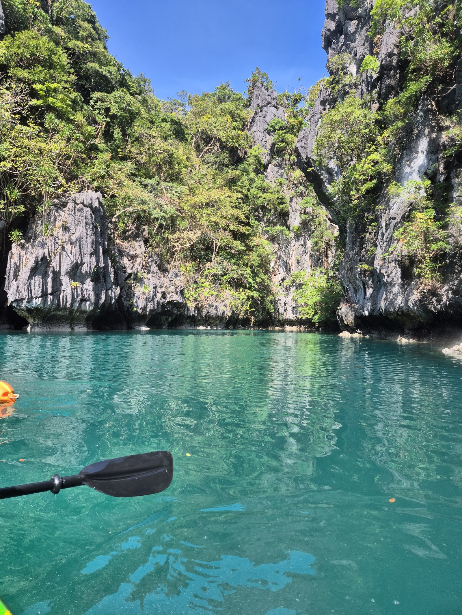 A kayak paddle extends into turquoise lagoon water surrounded by towering limestone cliffs covered in green vegetation.
