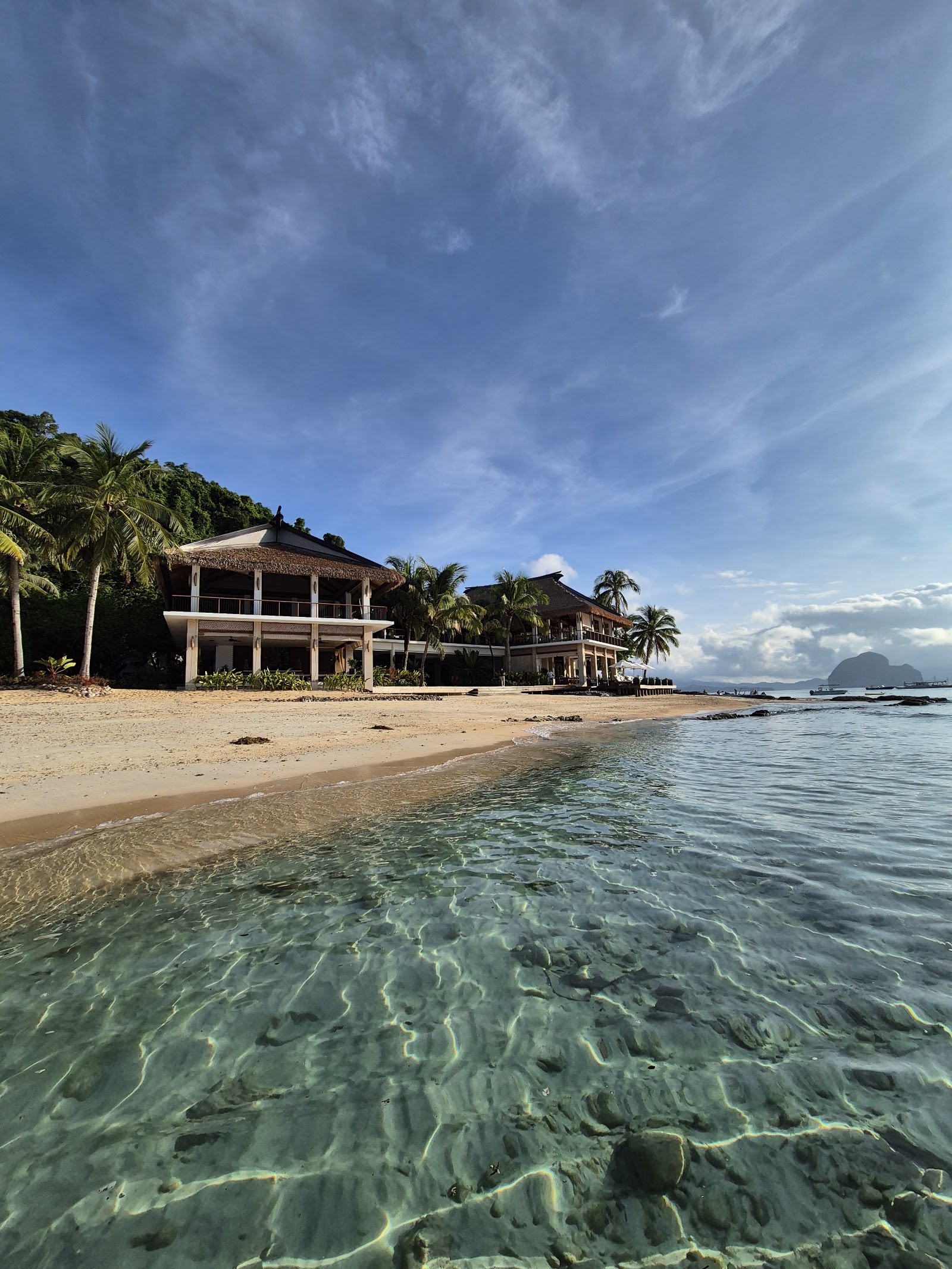 Clear shallow water laps a sandy beach in front of thatch-roofed beachfront buildings with palm trees and mountains visible across the bay.