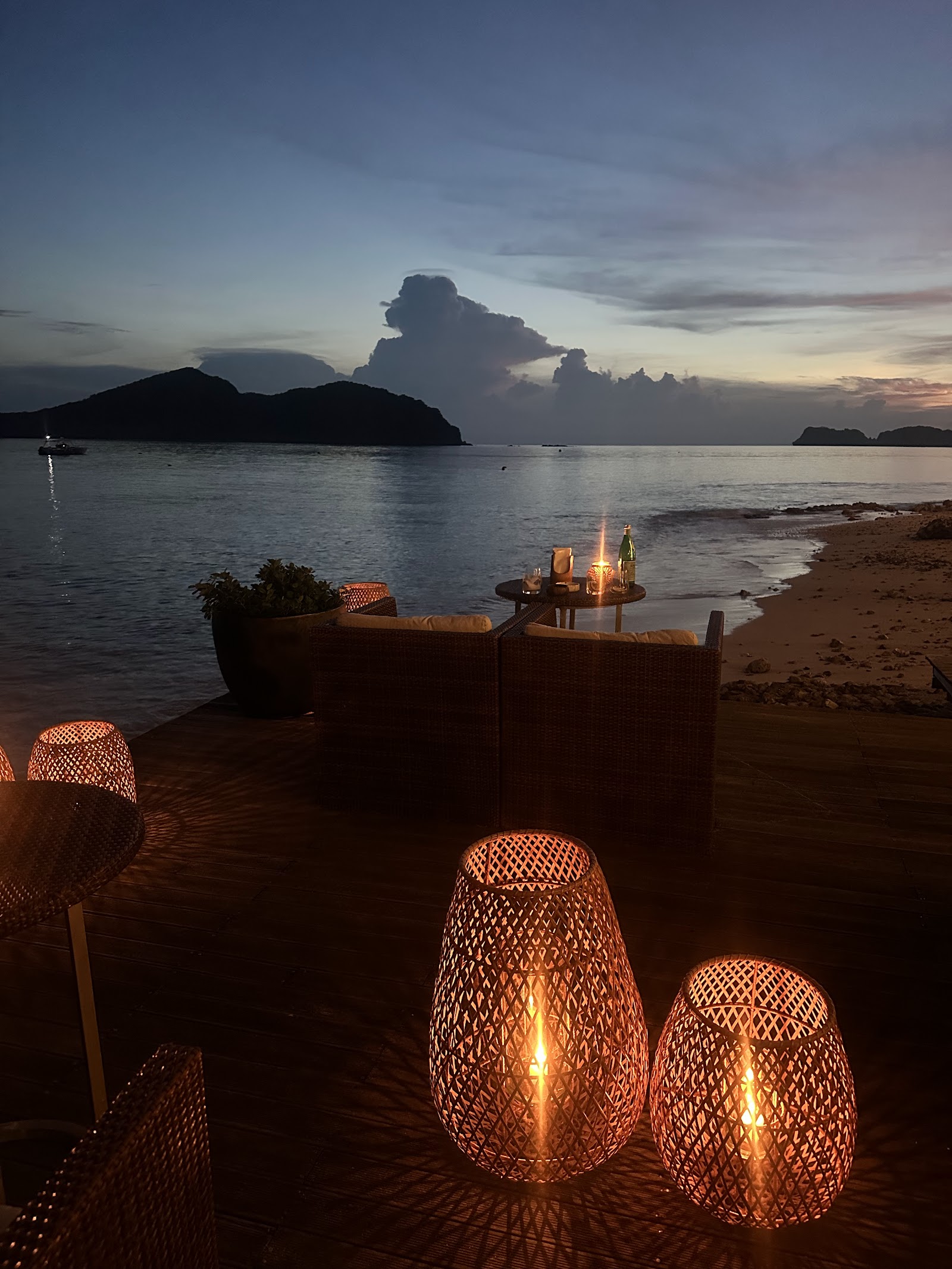 Beachfront dining setup at dusk with woven lanterns, rattan seating, and a table with drinks overlooking islands and calm waters.
