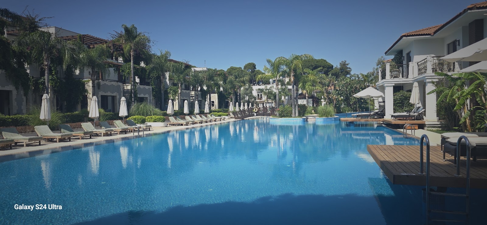 Large lagoon-style pool with bright blue water, white umbrellas lining the beach, a poolside bar pavilion, and landscaped grounds under clear sky at Ela Excellence Resort Belek.