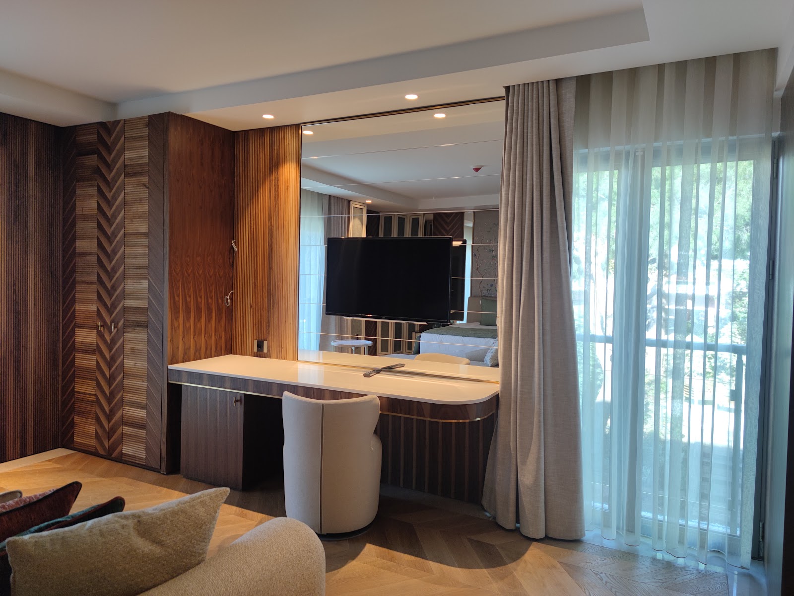 Resort suite with curved desk, patterned wood cabinets, flat-screen TV, and glass doors opening to a balcony overlooking the grounds.