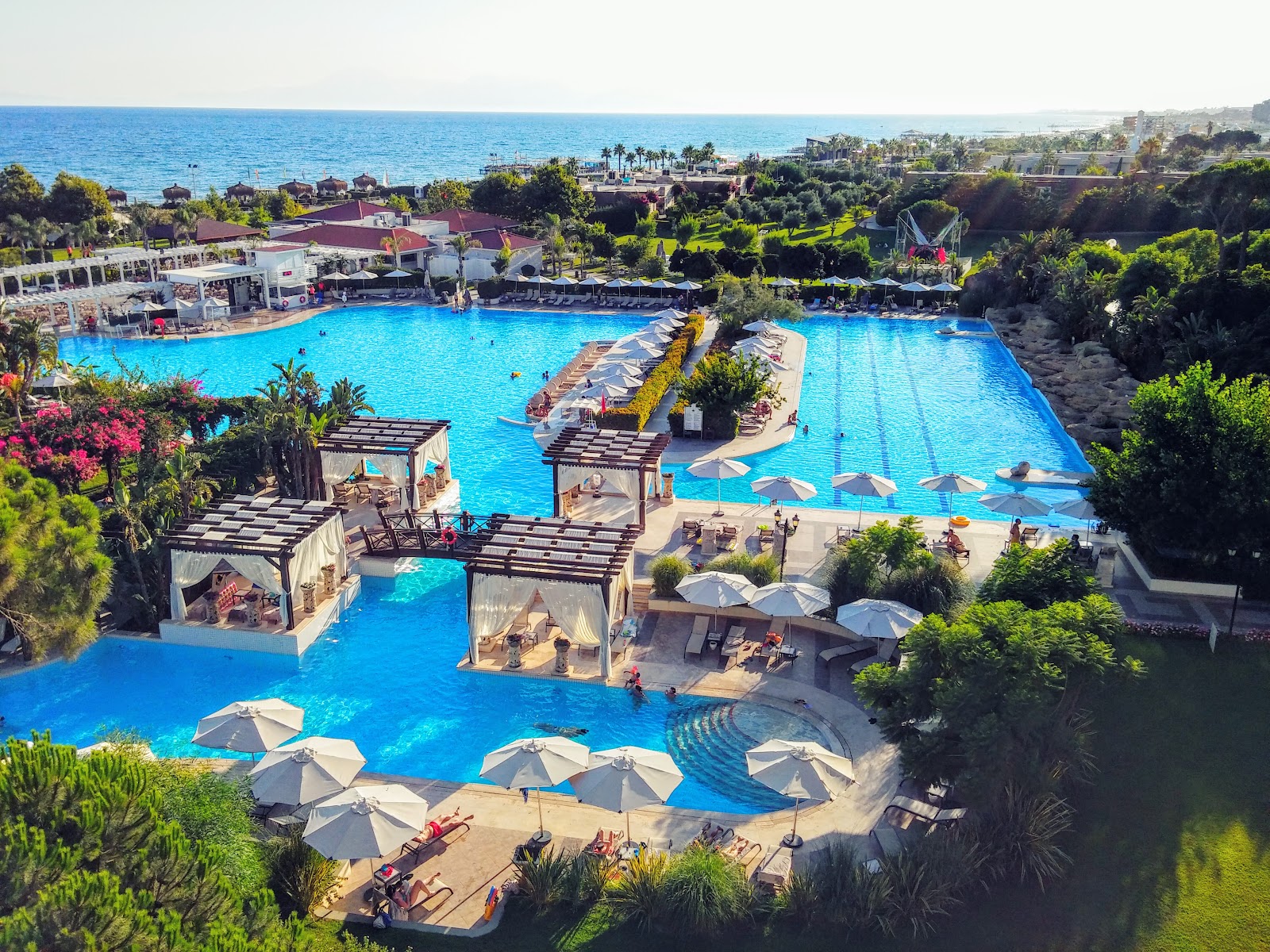 Aerial view of Ela Excellence Resort Belek's large turquoise pools with cabanas, white umbrellas, and the Mediterranean Sea beyond.