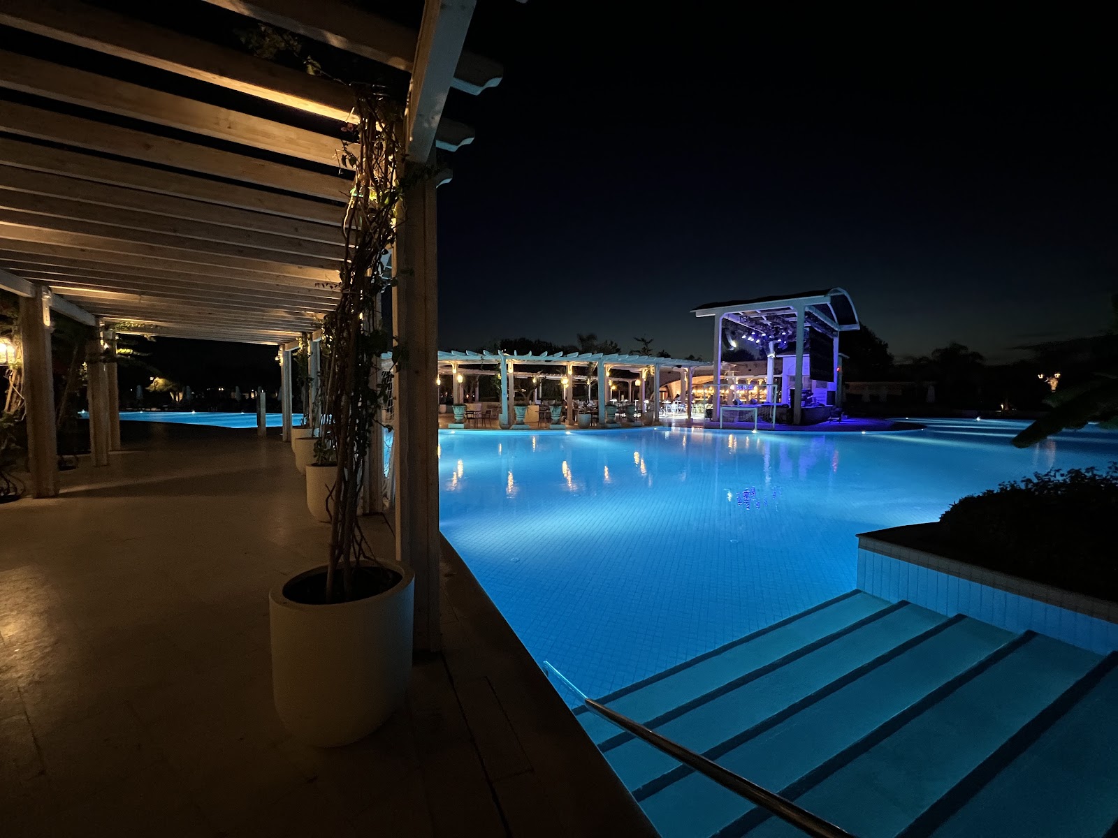 Nighttime view of the illuminated pool at Ela Excellence Resort Belek, with lit pergolas and bar pavilion reflected in the blue water.