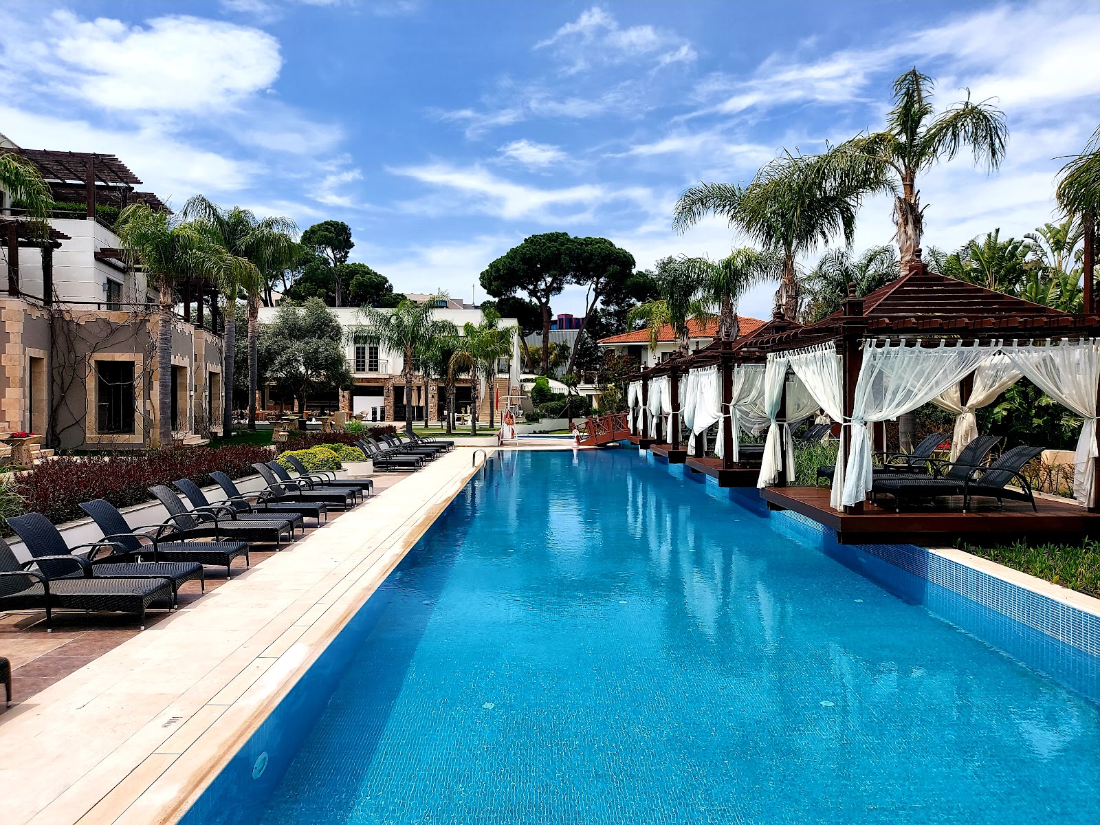 Rectangular swimming pool with loungers and cabanas lined on either side, palm trees and resort buildings in background under blue sky.