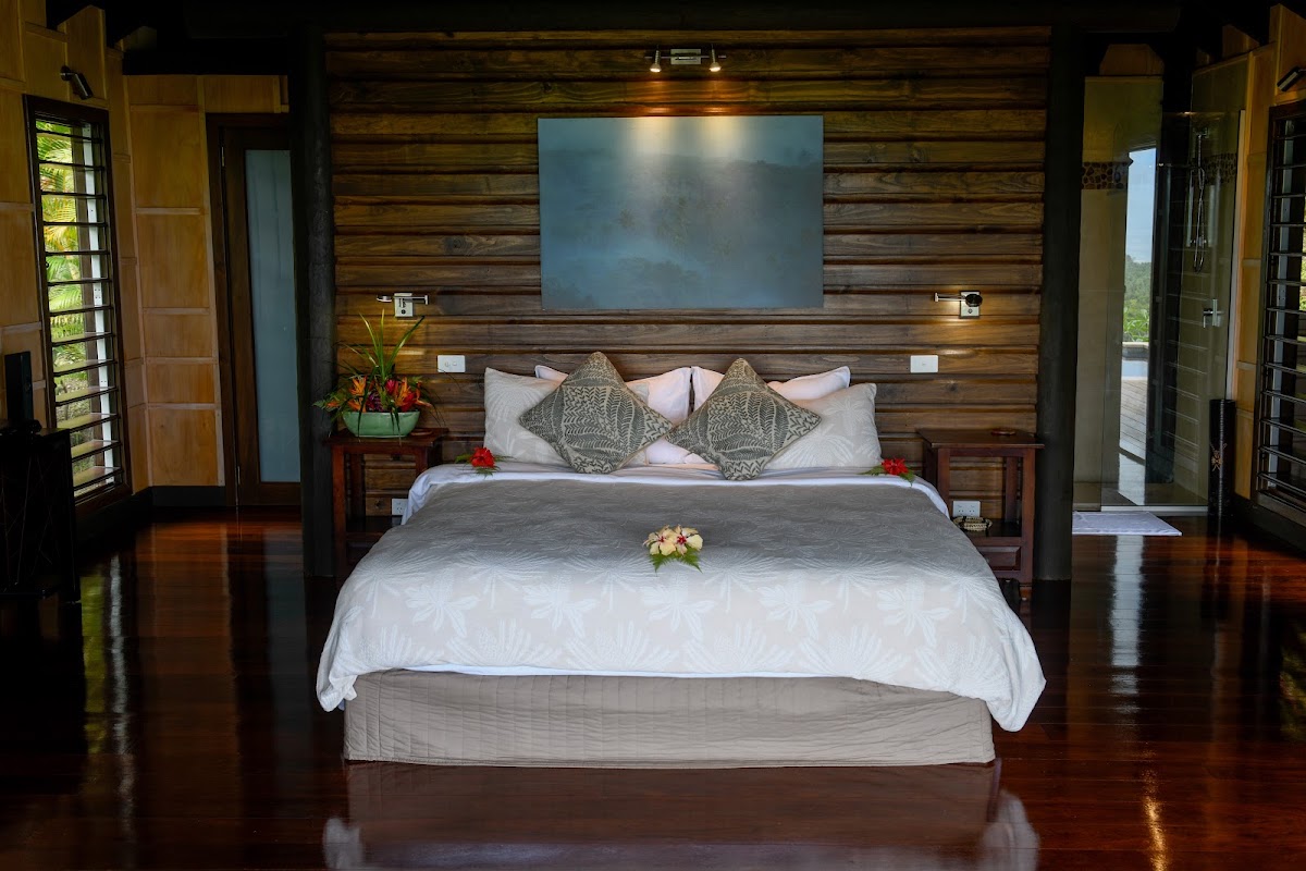Spacious bedroom with wooden log walls, king bed with white linens and decorative pillows, wall-mounted TV, and dark wood flooring with open doors to a terrace.